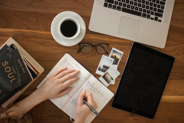 woman journaling on a desk next to her laptop, tablet, and books