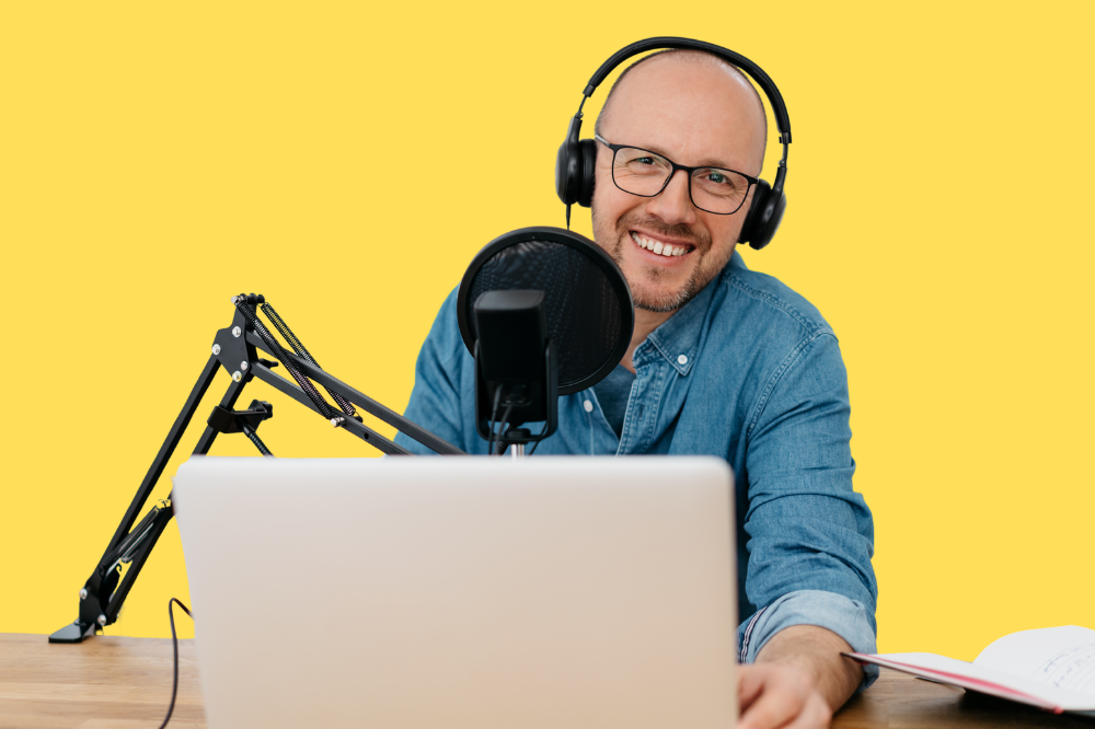Smiling podcaster with headphones and microphone sitting at a desk with a laptop
