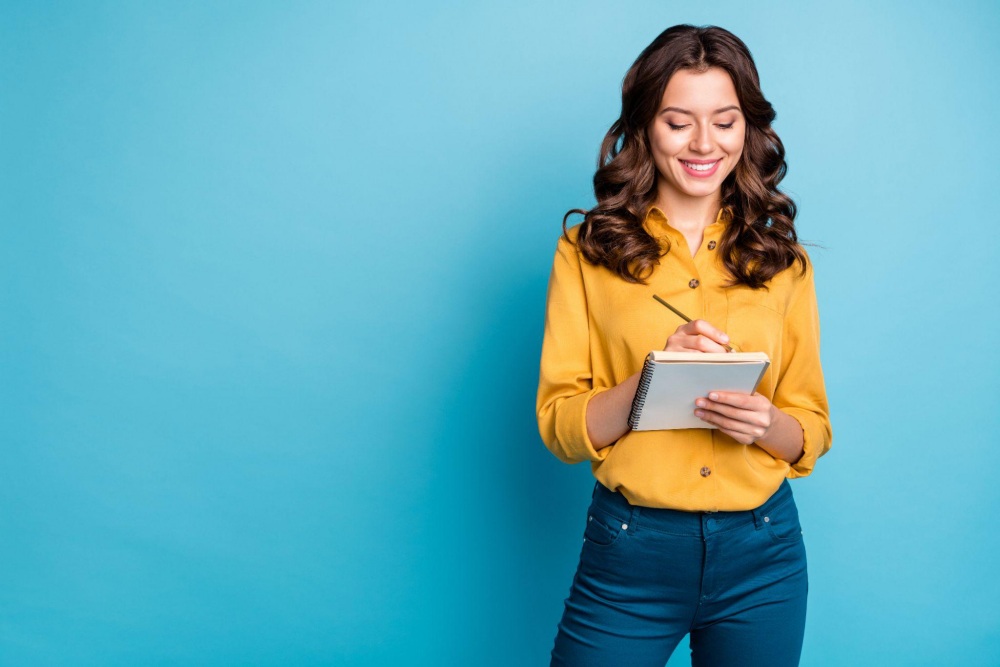 Smiling woman in yellow top writing in a notebook against a blue background