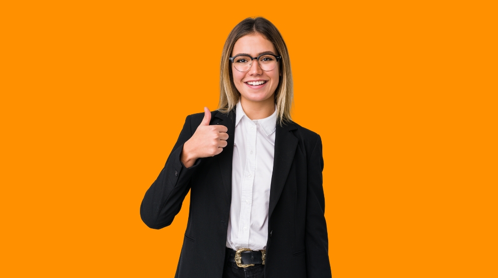 Smiling woman in blazer giving thumbs up on orange background
