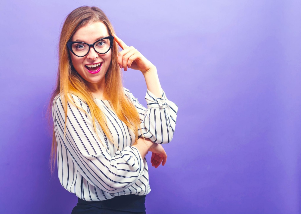Smiling woman with long hair and glasses pointing to her head in front of a purple background