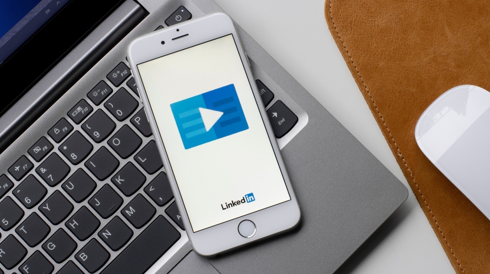 Smartphone on laptop keyboard showing LinkedIn video play button next to mouse