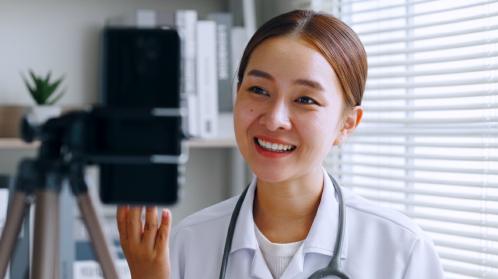 Smiling doctor in white coat waving near phone on tripod with bookshelves