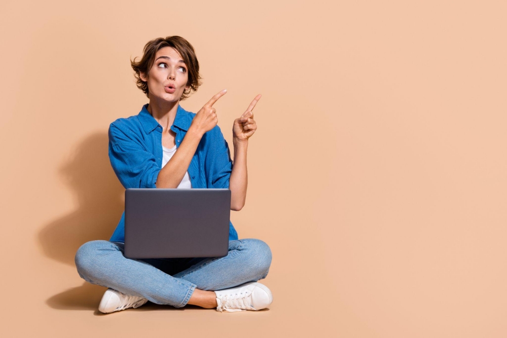 Woman in blue shirt and jeans sitting cross-legged with laptop, pointing upwards on beige background