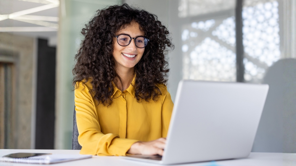 Smiling woman with curly hair in yellow shirt working on laptop