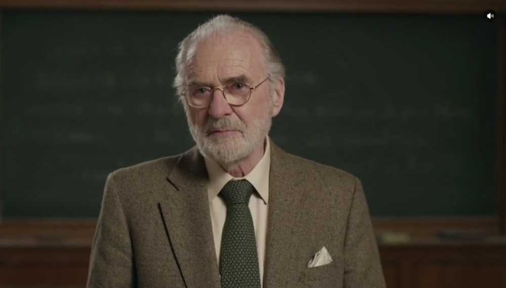 Elderly man with white hair and beard in suit and tie standing in front of chalkboard
