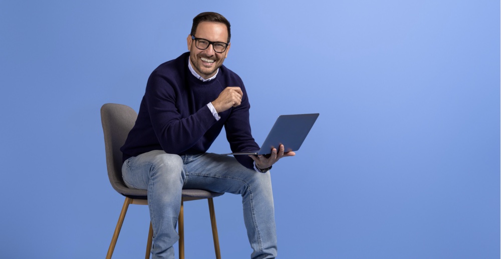 Smiling man with glasses holding laptop on chair.