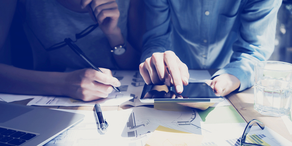 Two people working at a table top with documents and a tablet.