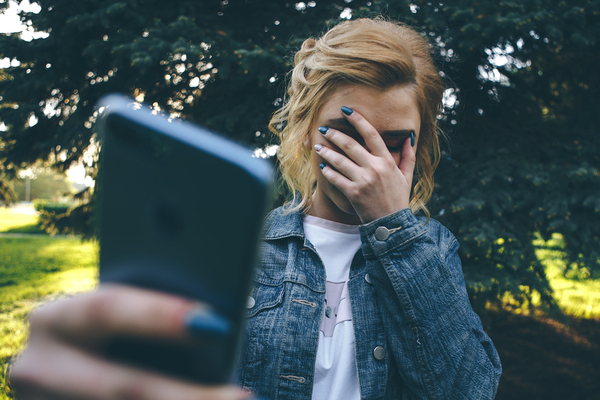Teen holding her phone in one hand and covering her face with the other hand.