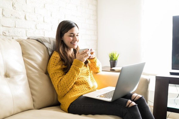 Woman sitting on a couch drinking a coffee and looking at a laptop.