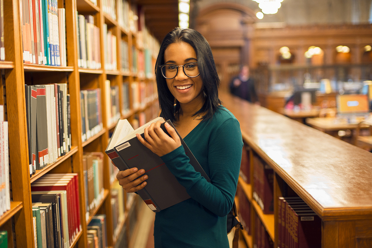 Woman in a library reading a book and smiling.