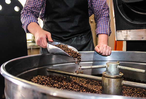 Person using a metal scoop to get coffee beans from a large mixer.