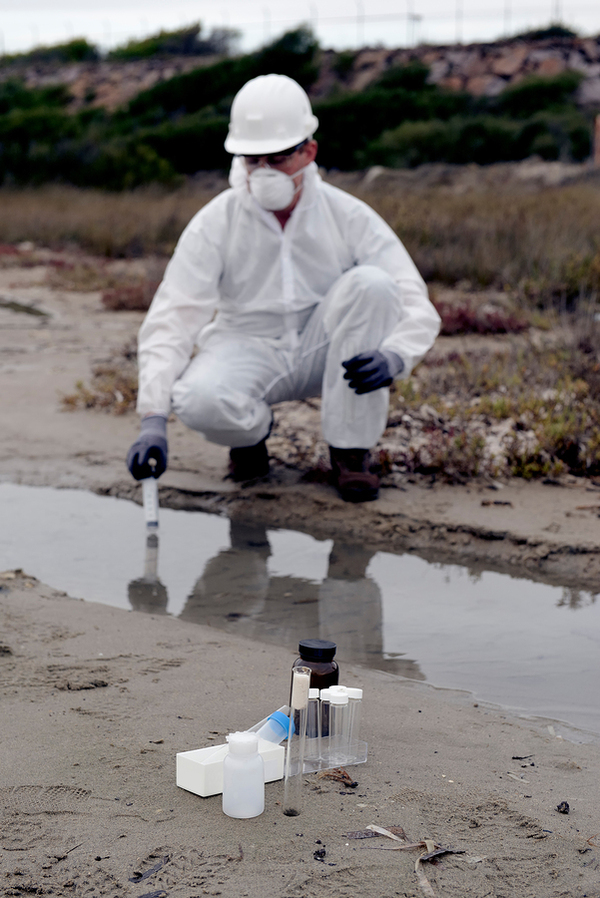 Man in a white jumpsuit, mask and hat taking a water sample.