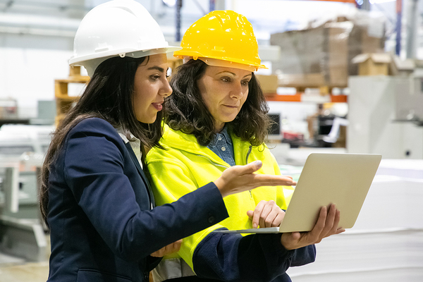 Two women wearing hard hats discussing information on a laptop.